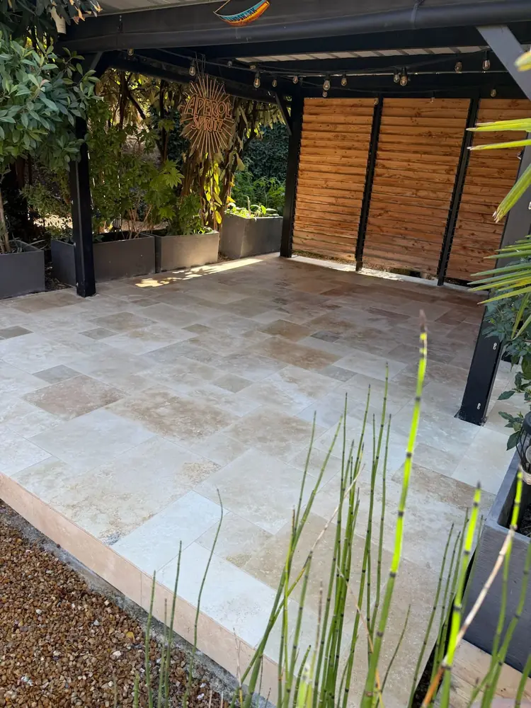 Terrasse travertin opus romain sous carport bois, vue d'angle avec jardinières et plantes