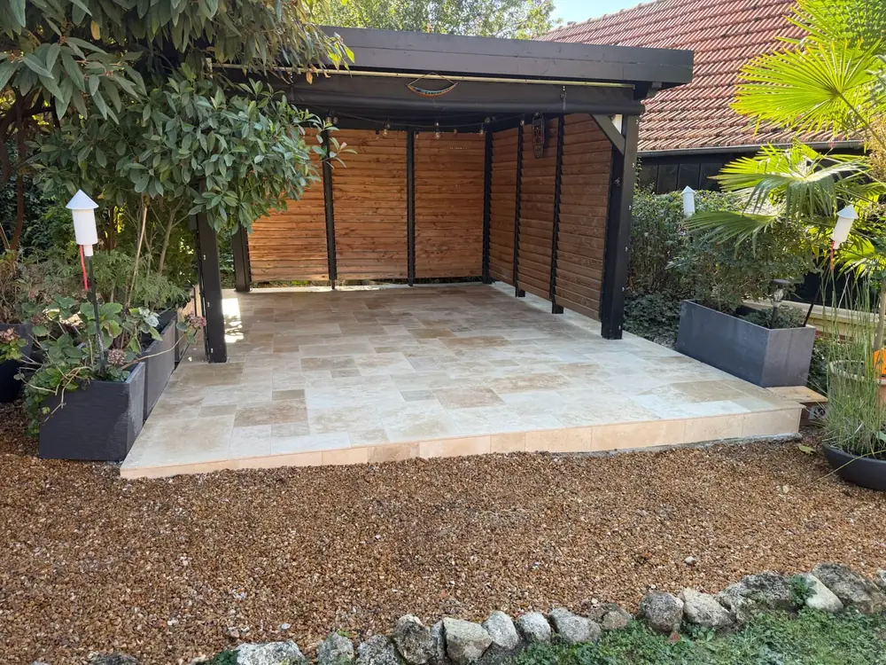 Terrasse en travertin beige posé en opus romain sous un carport en bois et acier noir, bordée de gravier